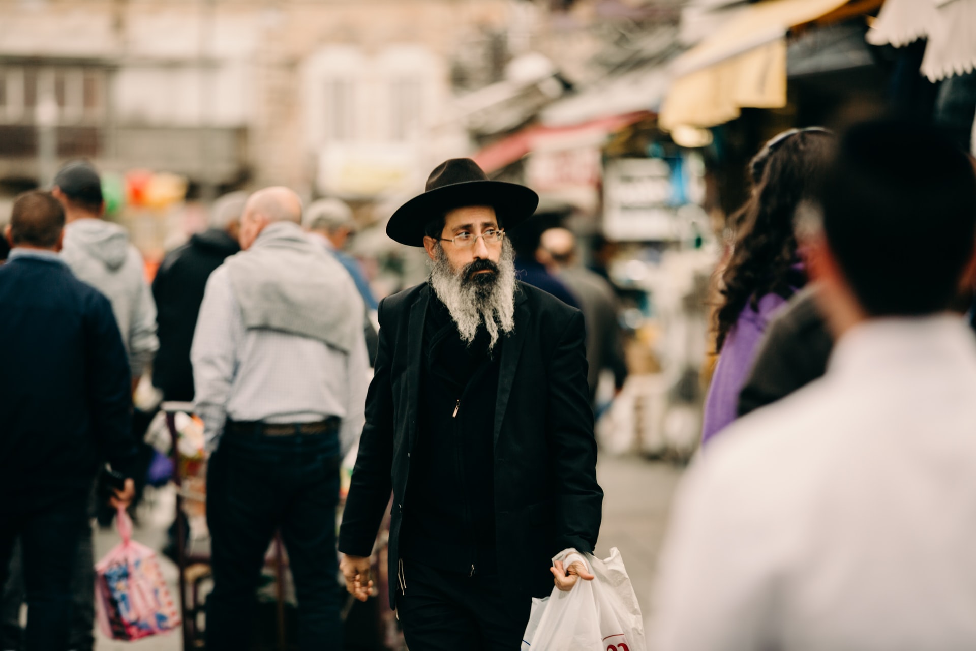 Várias pessoas caminhando nas ruas de Jerusalém, perto do mercado Mahne Yehuda Shuk, em foco um judeu ortodoxo vestido em roupas pretas segurando algumas sacolas nas mãos.
