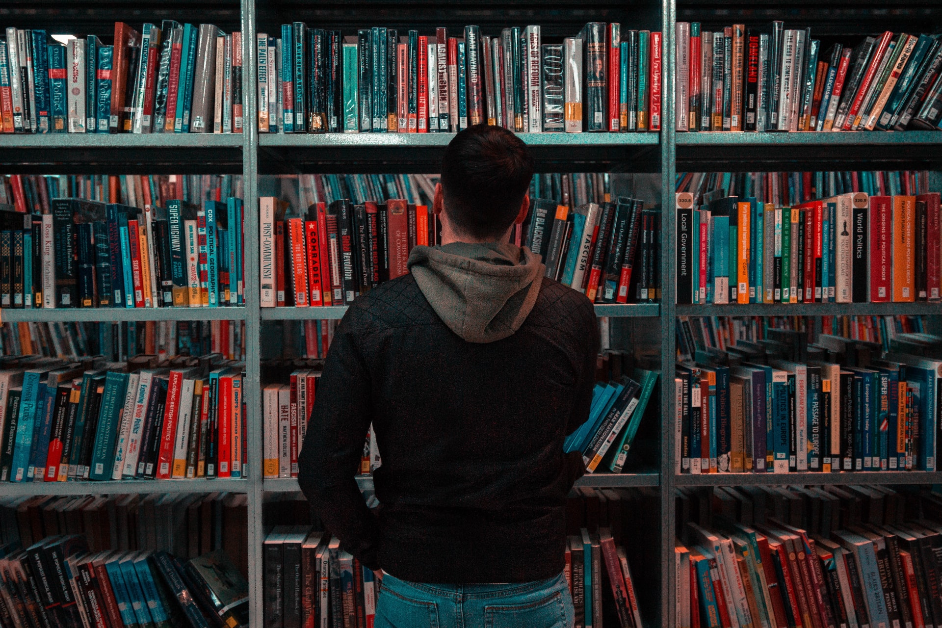 Homem em frente de uma estante cheia de livros, gesto corporal remetendo a dúvida, pensamento